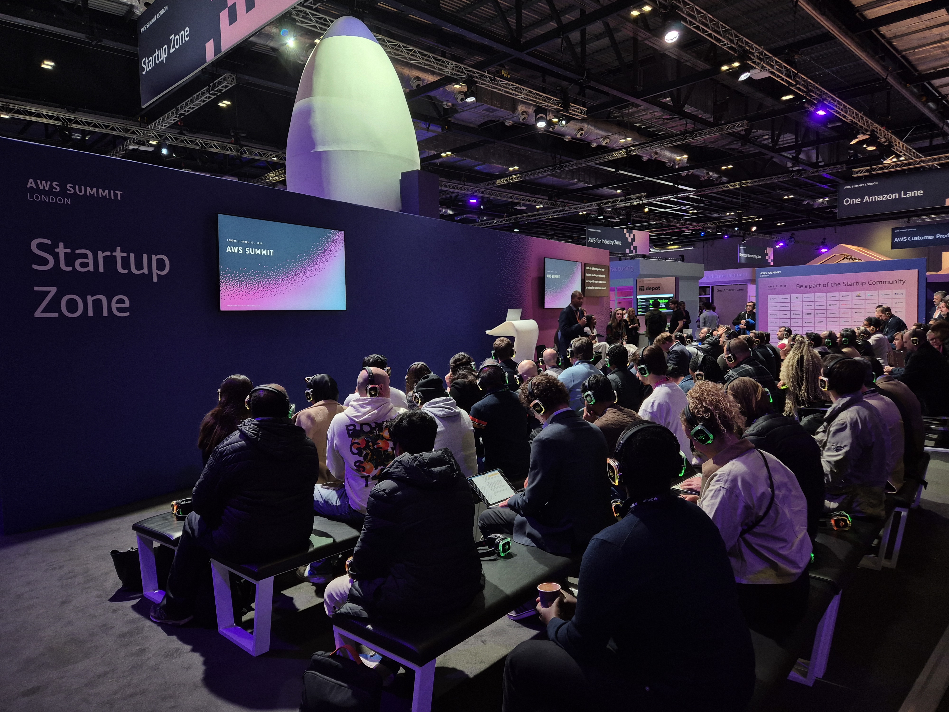 Crowded AWS Summit London 2026 startup zone with attendees wearing silent-disco style headphones during a presentation