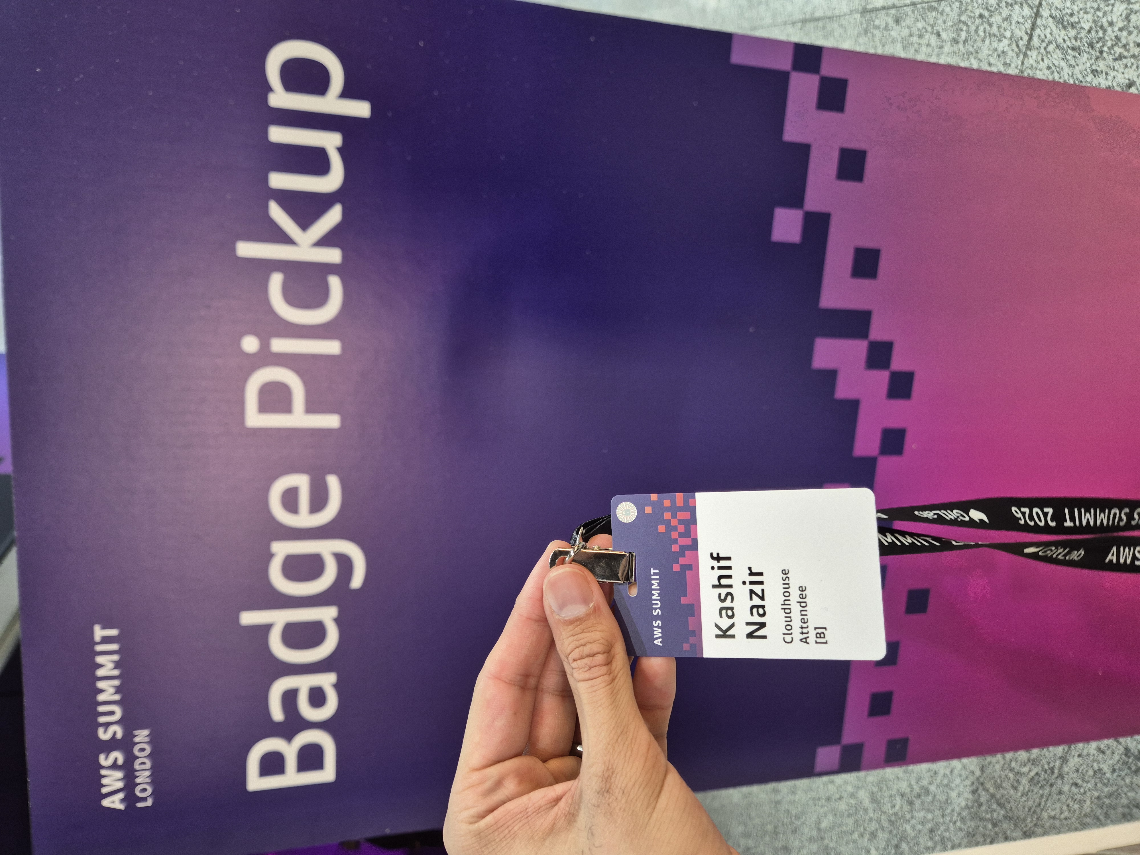 AWS Summit London 2026 attendee badge held in front of the Badge Pickup wall at ExCeL London