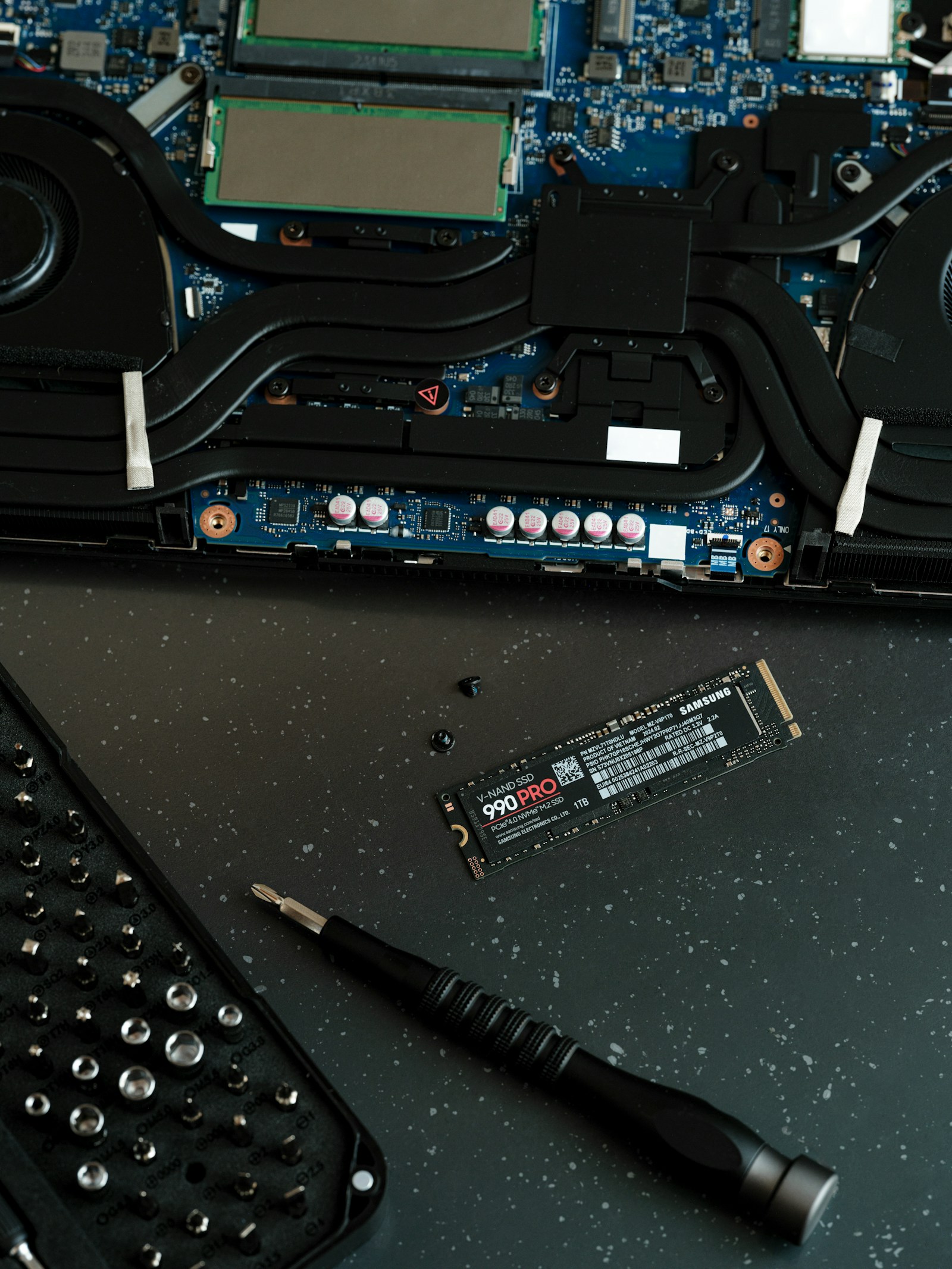 An opened laptop motherboard with cooling pipes, screws, tools, and a Samsung 990 Pro SSD beside it