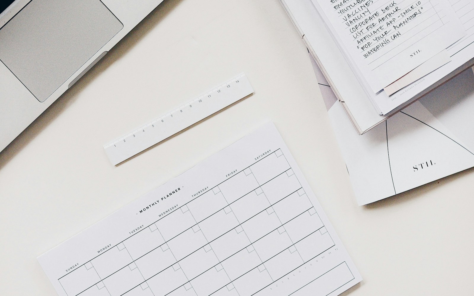 A laptop beside a notebook, planner, and handwritten task list on a white desk