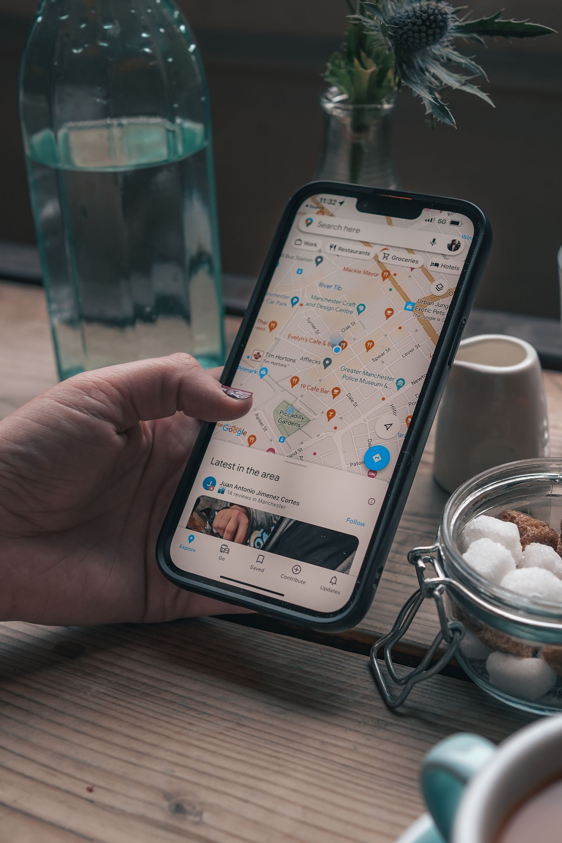 A hand holding a smartphone displaying Google Maps in a Manchester cafe